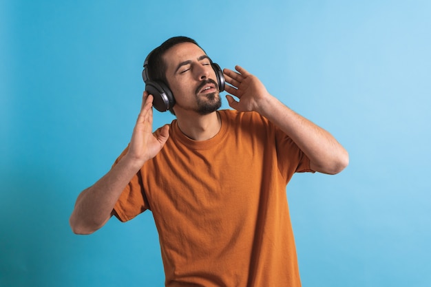 Young handsome man dancing and singing songs listening to music in headphones on a blue background