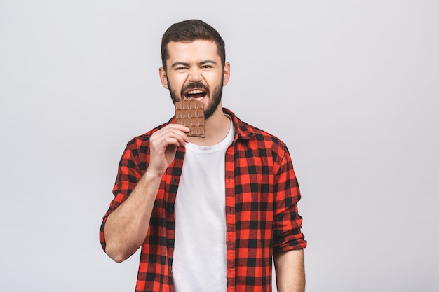 Young handsome man eating chocolate bar isolated against white background with a happy face standing and smiling with a confident smile showing teeth.