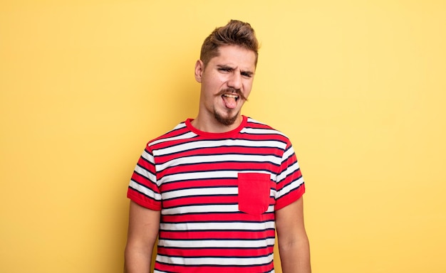 Young handsome man feeling disgusted and irritated, sticking tongue out, disliking something nasty and yucky. strange moustache concept