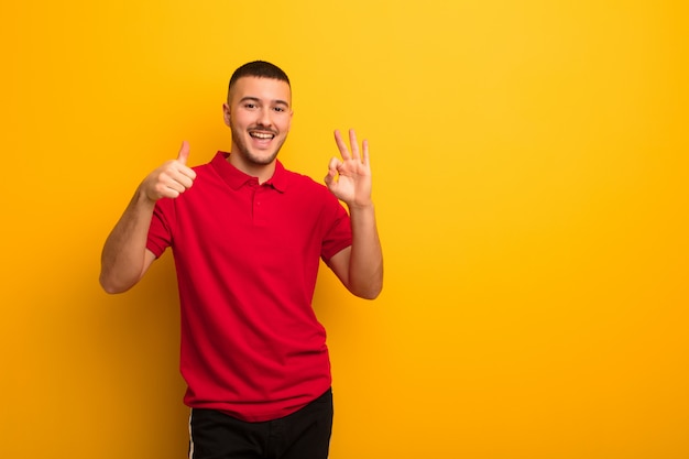 Young handsome man feeling happy, amazed, satisfied and surprised, showing okay and thumbs up gestures, smiling against flat