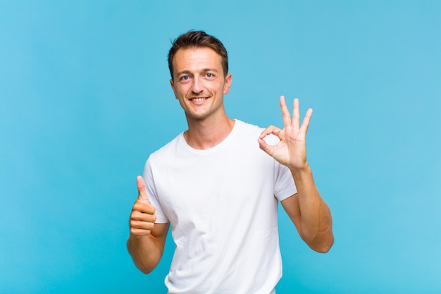 Young handsome man feeling happy, amazed, satisfied and surprised, showing okay and thumbs up gestures, smiling