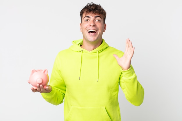 Young handsome man feeling happy and astonished at something unbelievable and holding a piggy bank