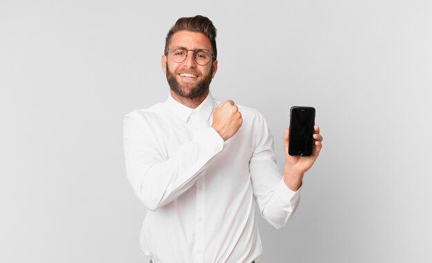 Young handsome man feeling happy and facing a challenge or celebrating and holding a cell