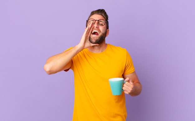 Young handsome man feeling happy,giving a big shout out with hands next to mouth. and holding a coffee mug