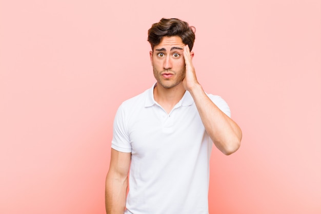 Young handsome man feeling shocked and astonished holding face to hand in disbelief with mouth wide open against pink background