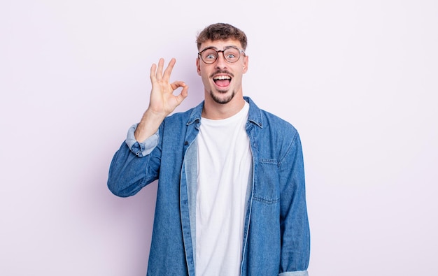 Young handsome man feeling successful and satisfied, smiling with mouth wide open, making okay sign with hand