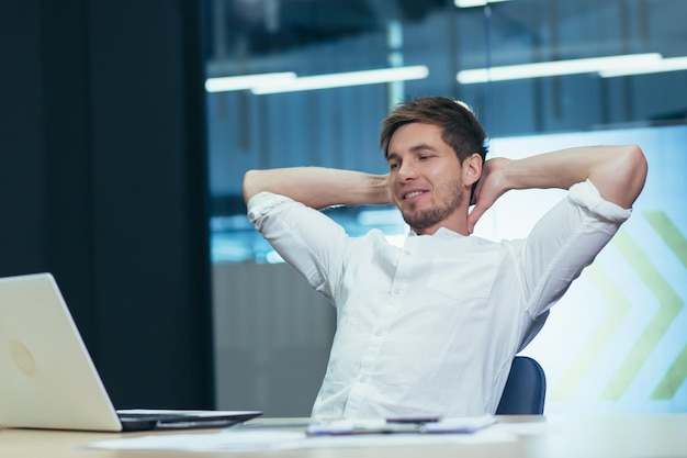 Young handsome man freelancer businessman manager in the office at the table resting doing work during a break throwing his hands behind his head smiling