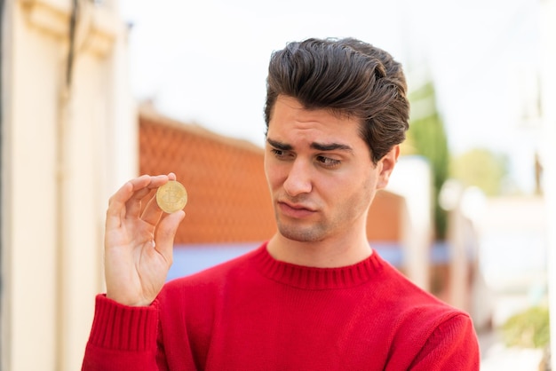 Photo young handsome man holding a bitcoin at outdoors with sad expression