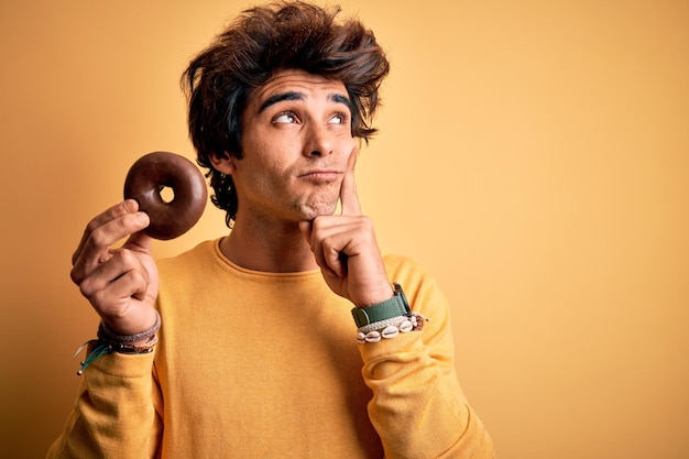 Young handsome man holding chocolate donut standing over isolated yellow background serious face thinking about question very confused idea