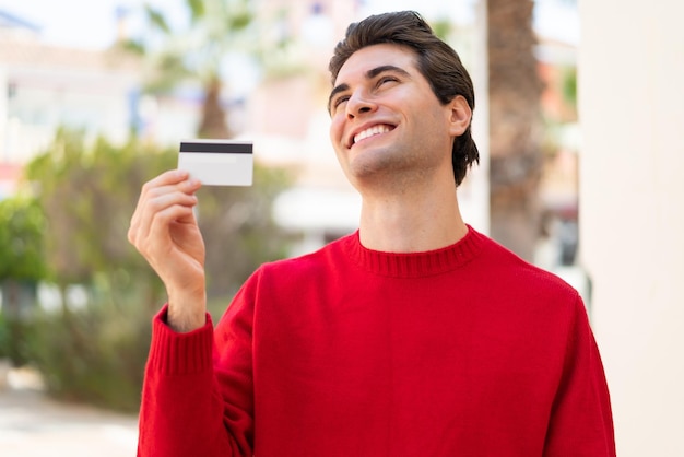 Young handsome man holding a credit card at outdoors looking up while smiling