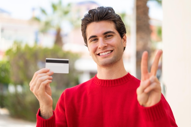 Young handsome man holding a credit card at outdoors smiling and showing victory sign