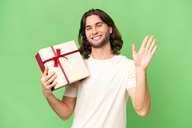 Young handsome man holding a gift over isolated background saluting with hand with happy expression