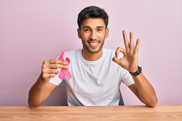 Young handsome man holding pink cancer ribbon doing ok sign with fingers, smiling friendly gesturing excellent symbol