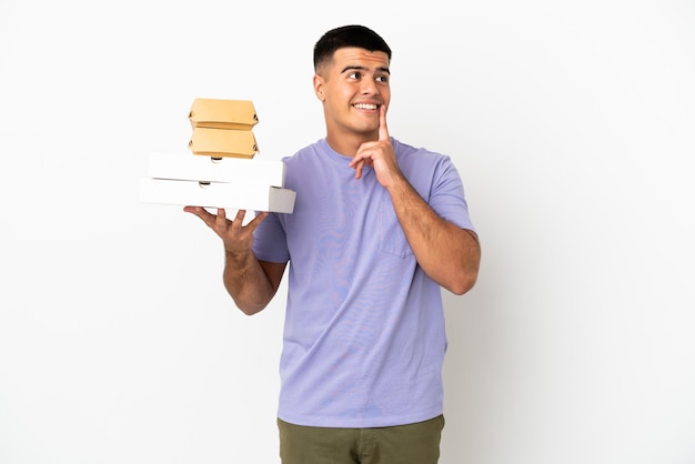 Young handsome man holding pizzas and burgers over isolated white wall thinking an idea while looking up