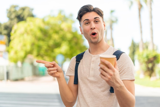 Young handsome man holding a take away coffee at outdoors surprised and pointing side