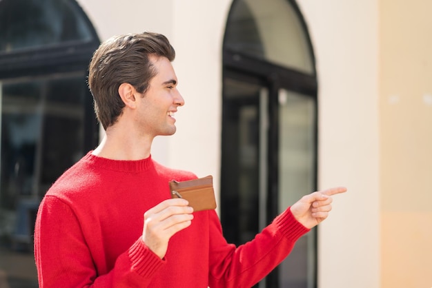 Young handsome man holding a wallet at outdoors pointing to the side to present a product