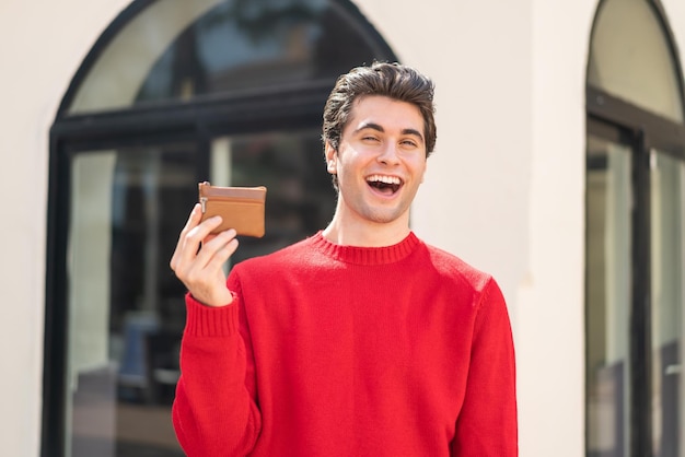 Young handsome man holding a wallet at outdoors with surprise and shocked facial expression