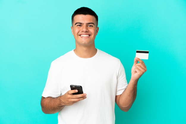 Young handsome man over isolated blue background buying with the mobile with a credit card