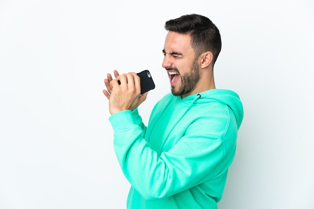 Young handsome man isolated on white wall using mobile phone and singing