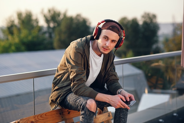 Young handsome man listening music in headphones and using phone on the rooftop of industrial building at the sunset.