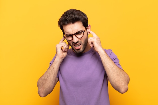 Young handsome man looking angry, stressed and annoyed, covering both ears to a deafening noise, sound or loud music