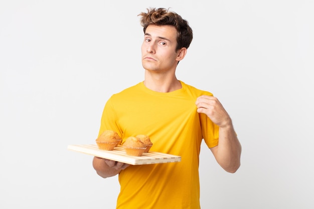 Young handsome man looking arrogant, successful, positive and proud holding a muffins tray