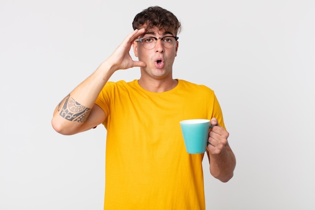 Young handsome man looking happy astonished and surprised and holding a coffe cup