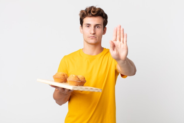 Young handsome man looking serious showing open palm making stop gesture holding a muffins tray