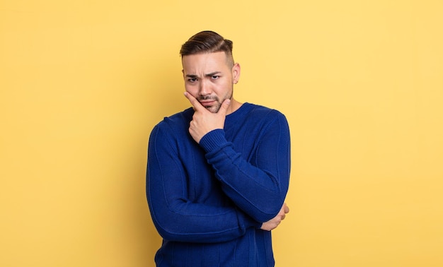 Young handsome man looking serious, thoughtful and distrustful, with one arm crossed and hand on chin, weighting options