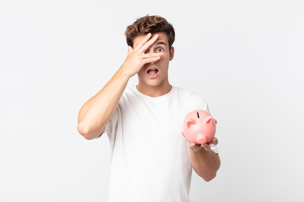 Young handsome man looking shocked, scared or terrified, covering face with hand and holding a piggy bank