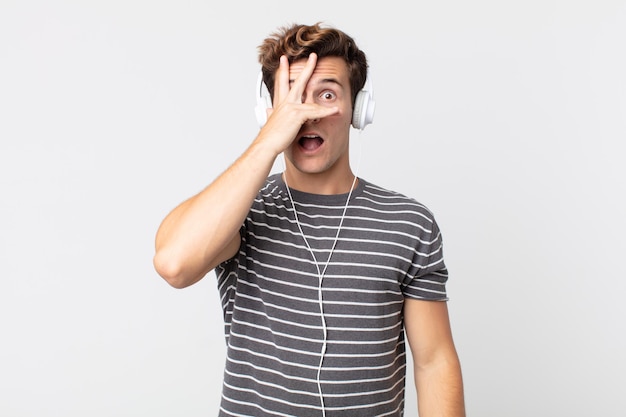 Photo young handsome man looking shocked, scared or terrified, covering face with hand. listening music concept