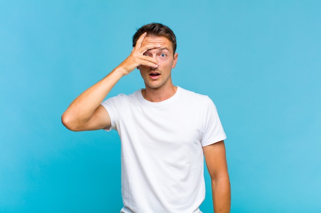 Young handsome man looking shocked, scared or terrified, covering face with hand and peeking between fingers