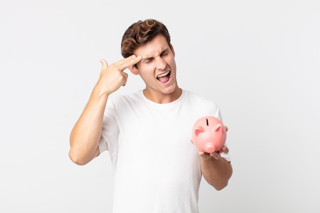 young handsome man looking unhappy and stressed, suicide gesture making gun sign and holding a piggy bank