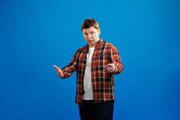 Young handsome man in shirt gesturing to you come here like invite you to come closer on blue studio background