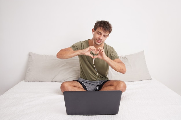 Young handsome man sitting on his bed on video call with his laptop computer, making heart symbol with his hands. Loved. Caucasian.