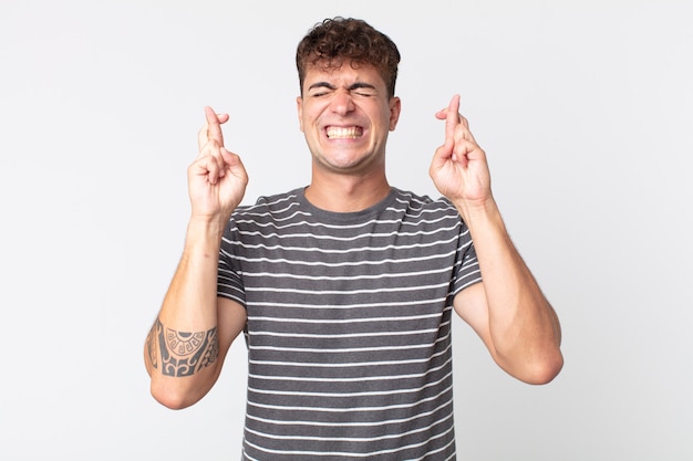 Young handsome man smiling and anxiously crossing both fingers, feeling worried and wishing or hoping for good luck