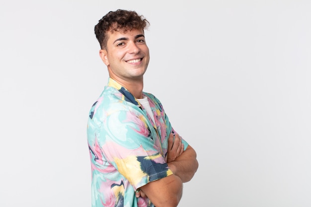 Young handsome man smiling to camera with crossed arms and a happy, confident, satisfied expression, lateral view