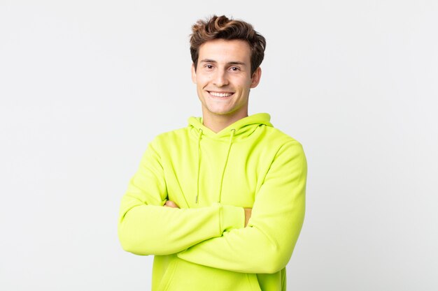 Young handsome man smiling to camera with crossed arms and a happy, confident, satisfied expression, lateral view