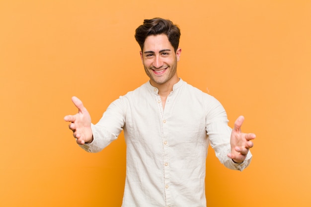 Young handsome man smiling cheerfully giving a warm, friendly, loving welcome hug, feeling happy and adorable against orange wall