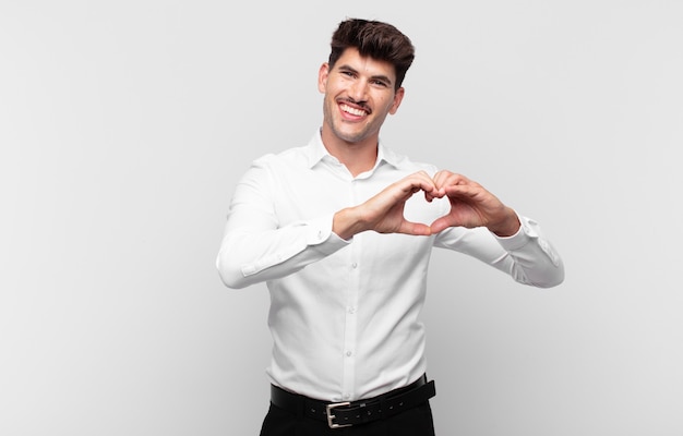 Young handsome man smiling and feeling happy, cute, romantic and in love, making heart shape with both hands