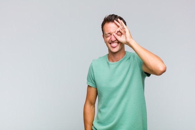 Young handsome man smiling happily with funny face, joking and looking through peephole, spying on secrets