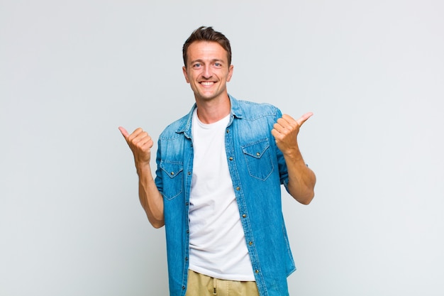 Young handsome man smiling joyfully and looking happy, feeling carefree and positive with both thumbs up