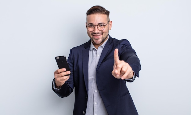 Young handsome man smiling and looking friendly, showing number one business and smartphone concept