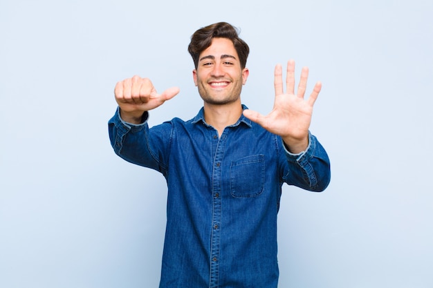 Young handsome man smiling and looking friendly, showing number six or sixth with hand forward, counting down against blue wall