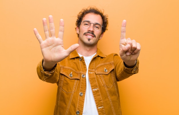 Young handsome man smiling and looking friendly, showing number six or sixth with hand forward, counting down against orange wall