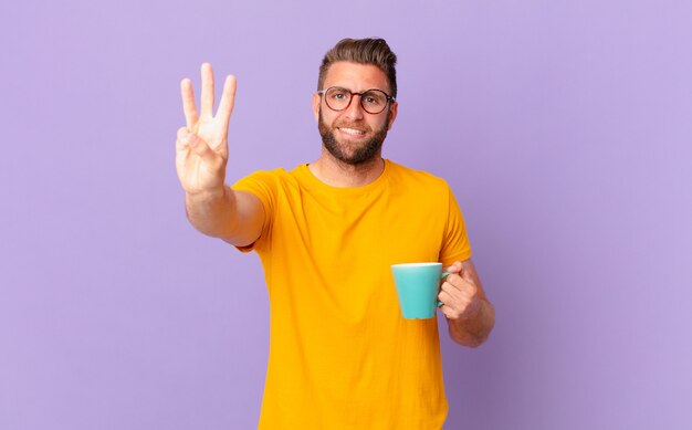 Young handsome man smiling and looking friendly, showing number three. and holding a coffee mug