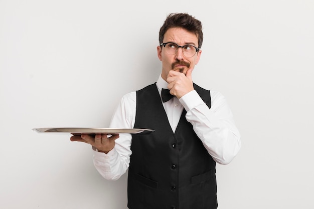 Young handsome man thinking feeling doubtful and confused waiter and tray concept