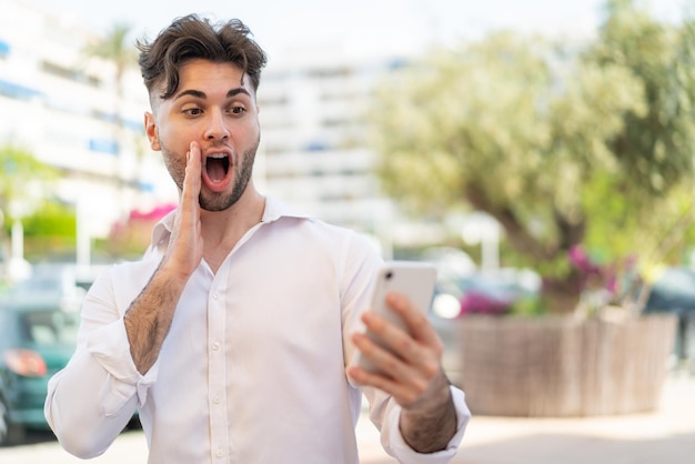 Young handsome man using mobile phone at outdoors with surprise and shocked facial expression