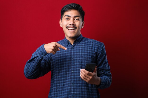 Young handsome man using smartphone wearing casual shirt isolated red background very happy pointing with hand and finger