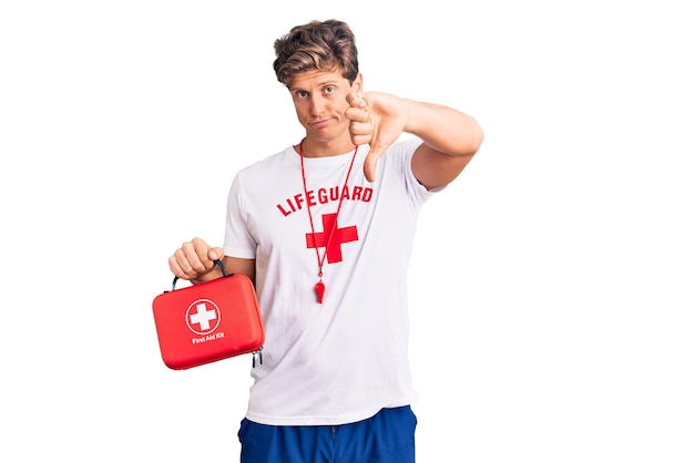 Young handsome man wearing lifeguard t shirt holding first aid kit with angry face negative sign showing dislike with thumbs down rejection concept